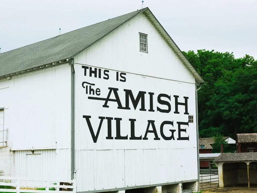 Amish Village Barn Sign A white barn with the words "THIS IS The AMISH VILLAGE" painted in large black letters on its side, set against a backdrop of green trees and a cloudy sky.