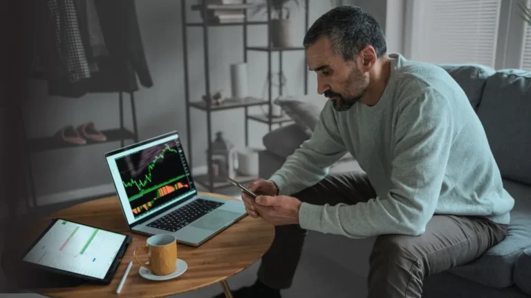 Man at home using his smartphone, potentially researching a reverse split calculator, with stock market charts visible on his nearby laptop and tablet.