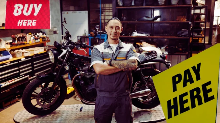 A friendly dealer stands in front of a motorcycle in a shop with signs that read "BUY HERE" and "PAY HERE," representing a buy here pay here motorcycles no credit check dealership